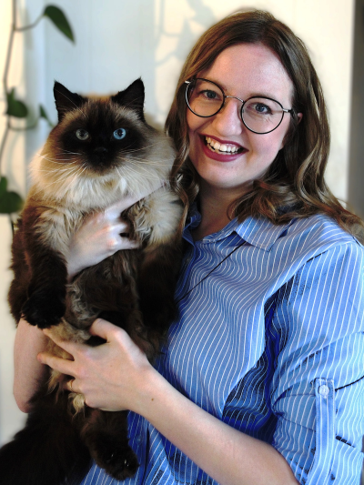 Chlie McMenamin with long brown hair and glasses, wearing a blue striped shirt, holding a fluffy dark brown and cream cat with bright blue eyes.
